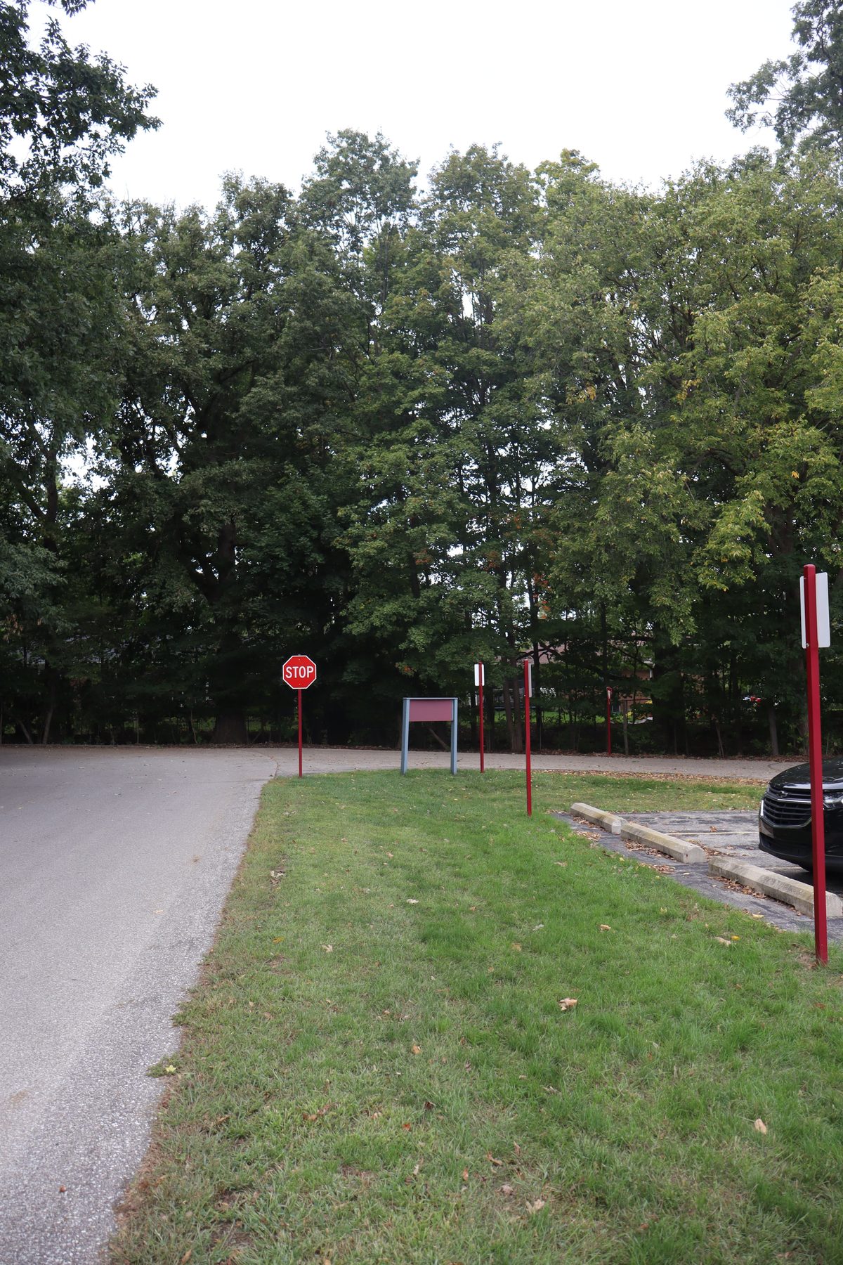 A stop sign and empty posts at the edge of a car park, trees behind